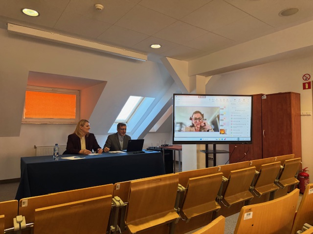 Dwie osoby siedzące za stołem kobieta, mężczyzna oraz kobieta na ekranie telewizora