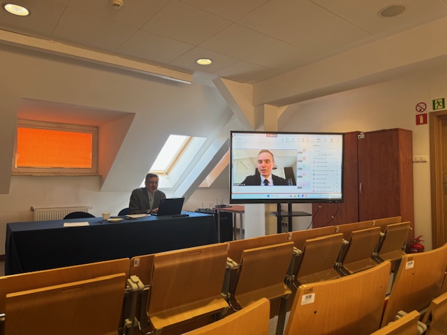 Siedzący mężczyzna za stołem oraz mężczyzna na ekranie telewizora